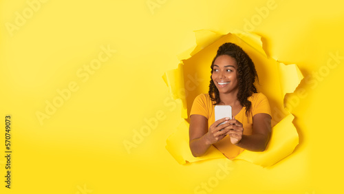 Happy african american woman with cellphone smiling and looking aside at free space through hole in torn paper, panorama