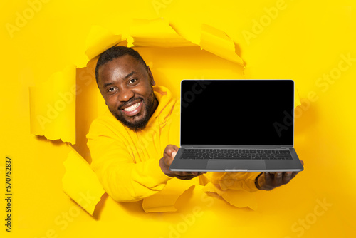 Excited black man holding laptop with empty blank screen showing device through yellow paper torn hole, mockup