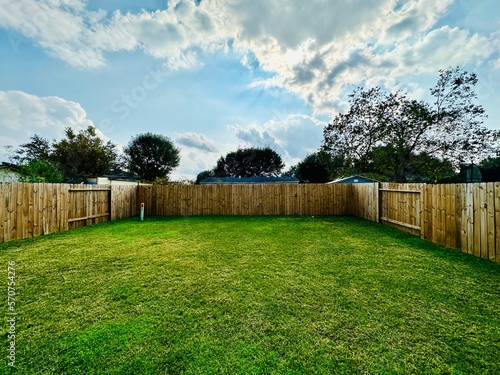 Large suburban backyard with a rustic wood fence, trimmed grass, and a concrete slab, set against neighboring homes under a clear blue sky.	