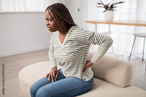 Black woman in pain sitting on couch, touching lower back