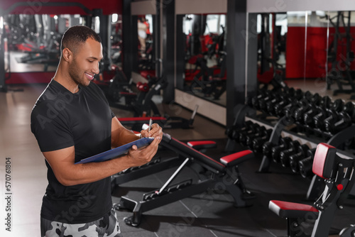 Happy trainer writing down workout plan in modern gym, space for text