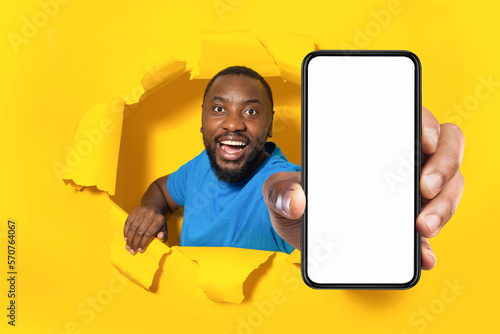 Happy african american man holding big cellphone with blank screen, through torn yellow paper hole, mockup