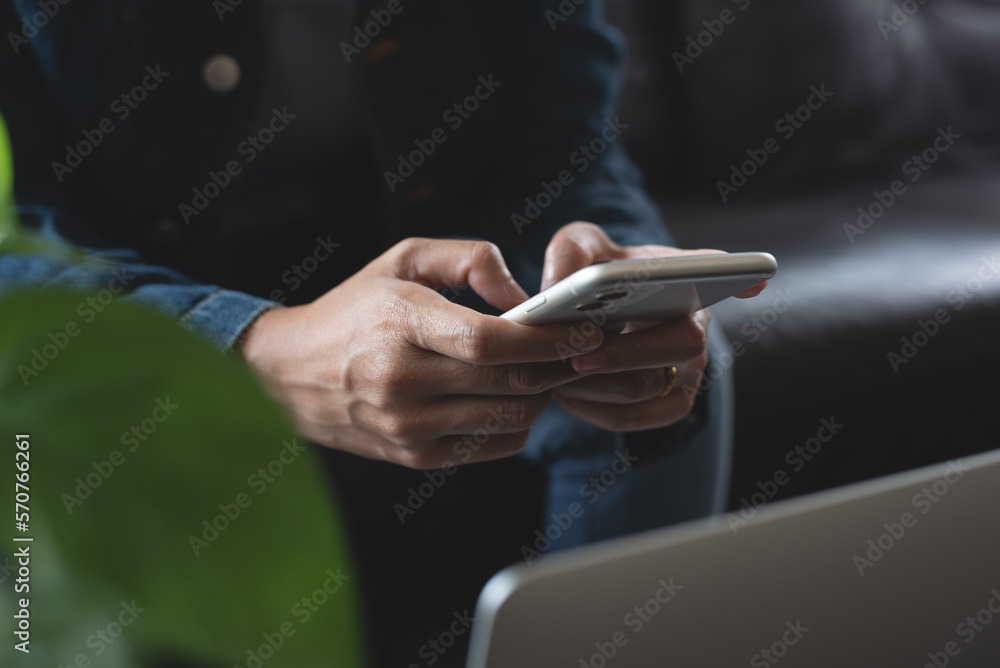 Woman sitting on sofa using mobile phone, working on laptop computer at home, close up