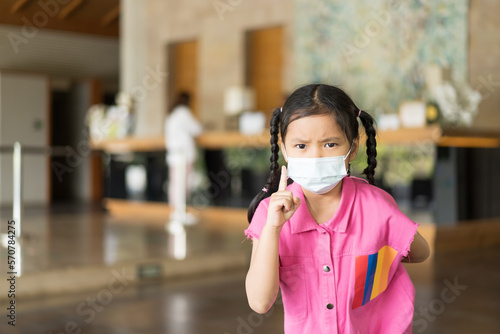 Asian child or kid girl wearing white face mask to serious pointing finger for close mouth nose or flu cough sick to protect PM2.5 dust or covid-19 coronavirus at hotel reception on holiday travel
