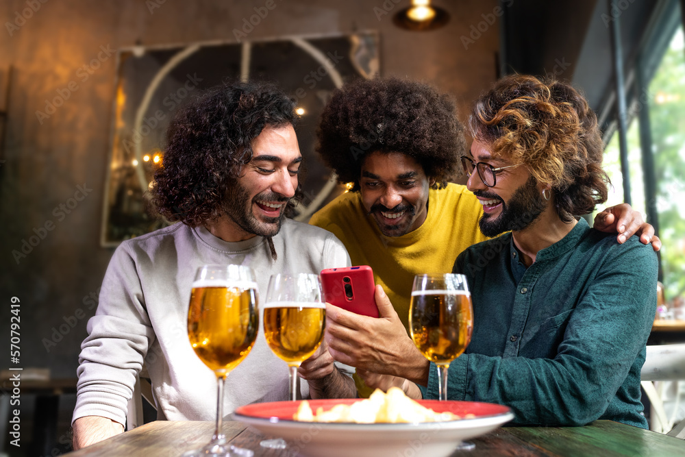 Young man showing to male friends mobile phone while having drinks together in pub. Young man showing to male friends mobile phone while having drinks together in pub.