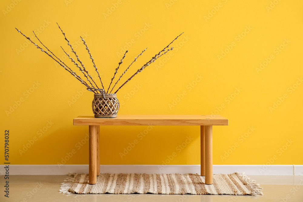 Vase with willow branches on wooden bench near yellow wall Vase with willow branches on wooden bench near yellow wall