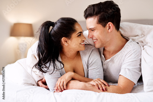 Happy, love and interracial couple in bed together in home holding hands at night. Smile, morning and marriage of an Indian woman and man with care and relax happiness in a bedroom in a house