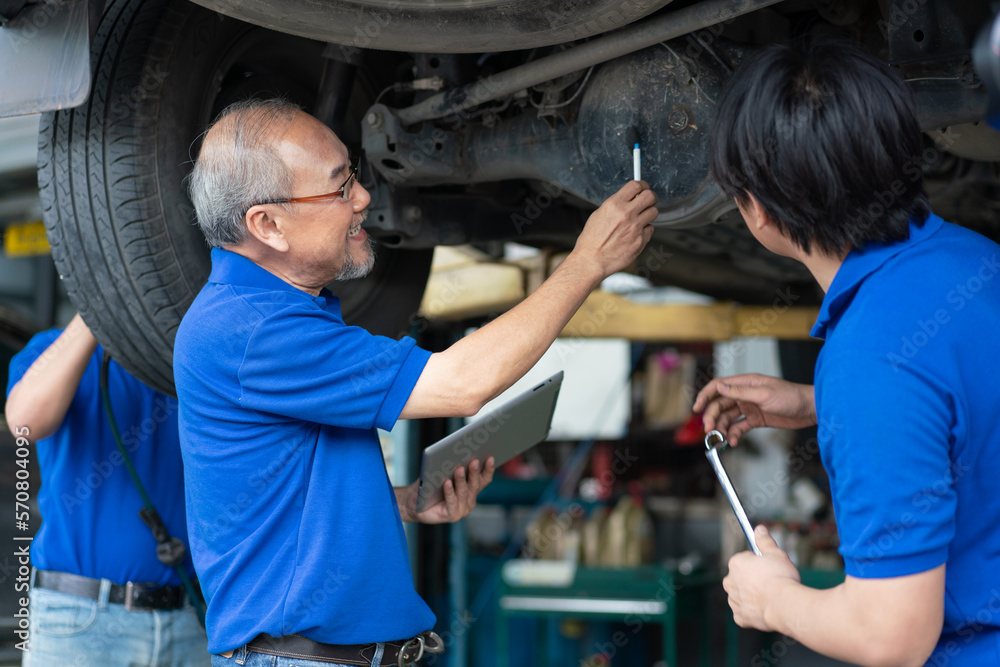 Senior Asian engineer teaching worker repair and maintenance car rear axle of the car. Group technician in uniform checking and servicing vehicle in garage. Senior Asian engineer teaching worker repair and maintenance car rear axle of the car. Group technician in uniform checking and servicing vehicle in garage.