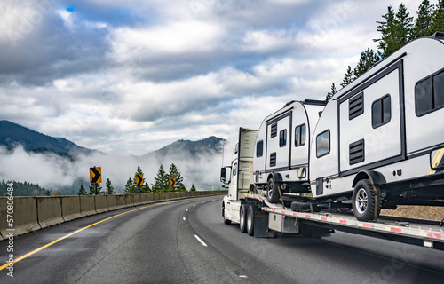 Long haul high cab white big rig semi truck transporting campers trucks on the step down semi trailer driving on the winding highway road with fog on the forest and mountain