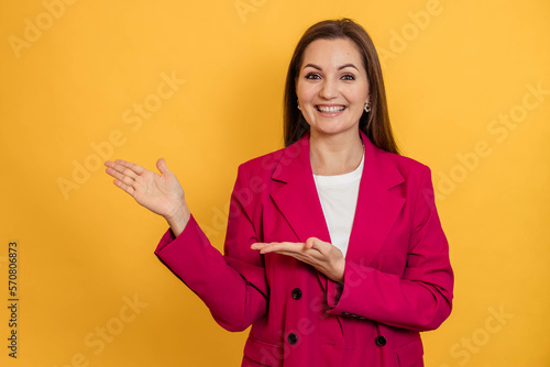 Portrait of a young girl in a pink jacket on a yellow background, pointing with both hands to the side © Natalia