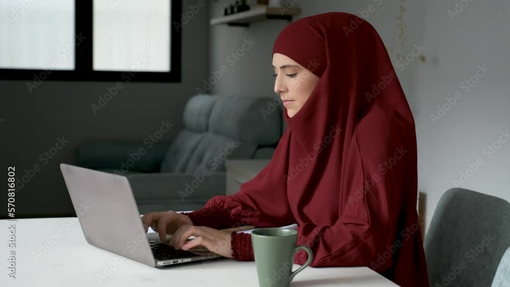 Muslim young woman in hijab on a phone call while working in a laptop at home. Muslim young woman in hijab on a phone call while working in a laptop at home.