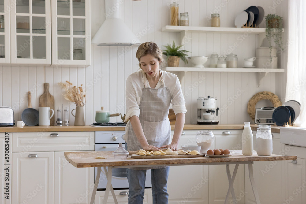 Calm pretty young baker woman in apron preparing pastry, bakery food for homemade dessert, pies, dumplings, rolling dough at table with flour, eggs, enjoying domestic activity