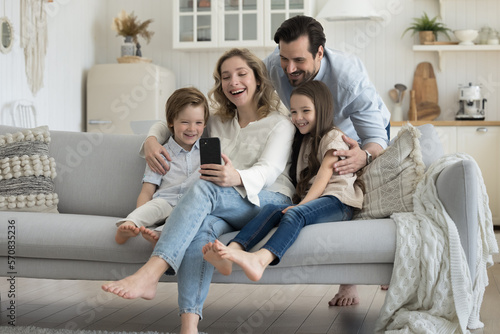 Happy millennial blogger parents hugging two little kids on sofa, shooting footage on smartphone, taking family selfie for social media, enjoying talk on video call, wireless online communication