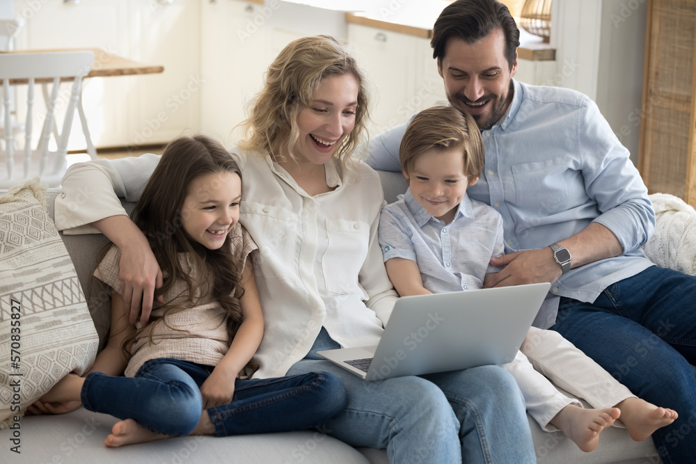Excited parents and two happy little kids using laptop at home for Internet communication, wireless online connection, relaxing on sofa together, watching movie, smiling, laughing Excited parents and two happy little kids using laptop at home for Internet communication, wireless online connection, relaxing on sofa together, watching movie, smiling, laughing