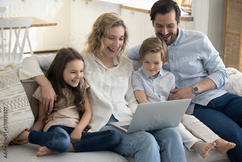 Excited parents and two happy little kids using laptop at home for Internet communication, wireless online connection, relaxing on sofa together, watching movie, smiling, laughing
