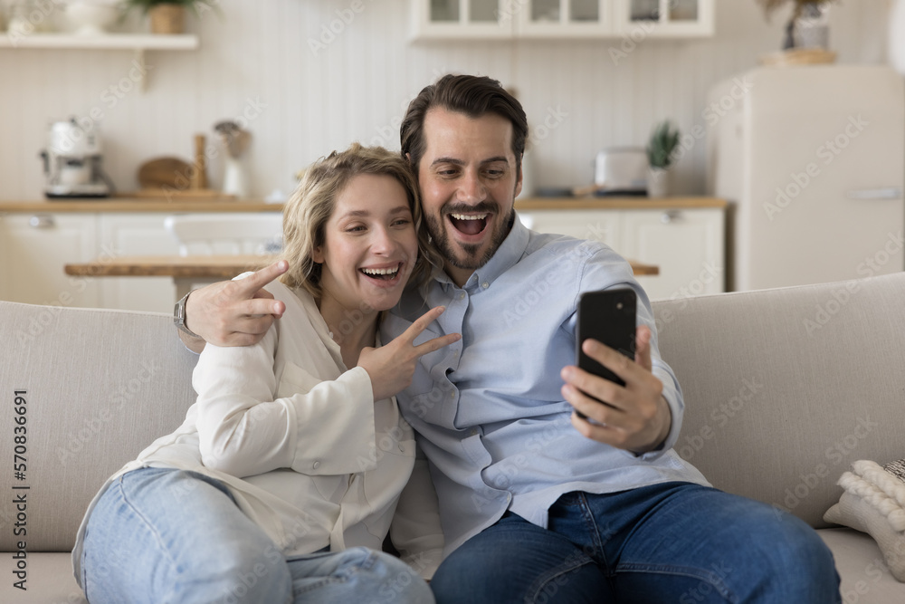 Fototapeta premium Cheerful positive young dating couple in love posing for selfie, taking self video, hugging on home sofa, showing Victory fingers gesture at smartphone, smiling, laughing, having fun