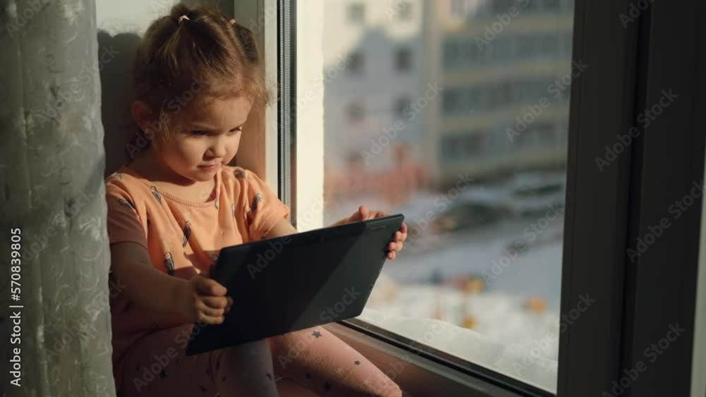 Child girl age 2 years old uses a tablet while sitting on the windowsill at home.Education at home. City life. Sunny morning. Child girl age 2 years old uses a tablet while sitting on the windowsill at home.Education at home. City life. Sunny morning.