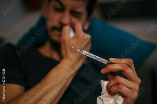 A sick Caucasian man having a fever, measuring his body temperature with a thermometer, touching his forehead in bed. Focus on a thermometer.