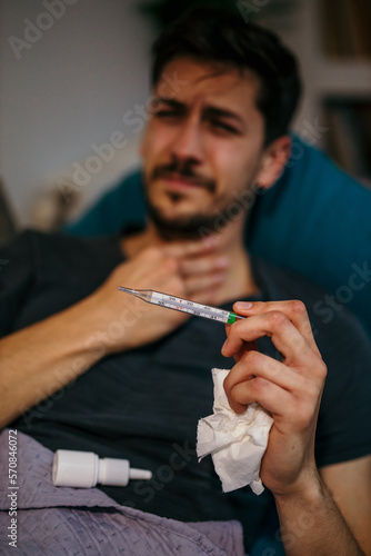 Man with a sore throat. The young Caucasian man sitting at home on a sofa  feeling sick  having a sore throat  and measuring his body temperature.