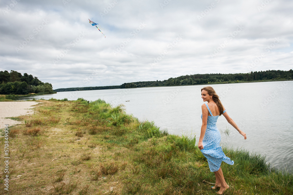 A beautiful slender smiling girl with long blond hair, in a blue summer dress, spins and walks along the river bank, against the backdrop of a picturesque landscape. A beautiful slender smiling girl with long blond hair, in a blue summer dress, spins and walks along the river bank, against the backdrop of a picturesque landscape.