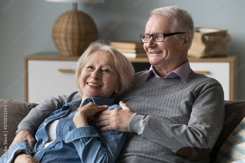 Happy cheerful elderly couple resting on sofa at home, hugging with love, enjoying leisure, retirement. Senior husband and wife relaxing on couch, talking, smiling, laughing Happy cheerful elderly couple resting on sofa at home, hugging with love, enjoying leisure, retirement. Senior husband and wife relaxing on couch, talking, smiling, laughing