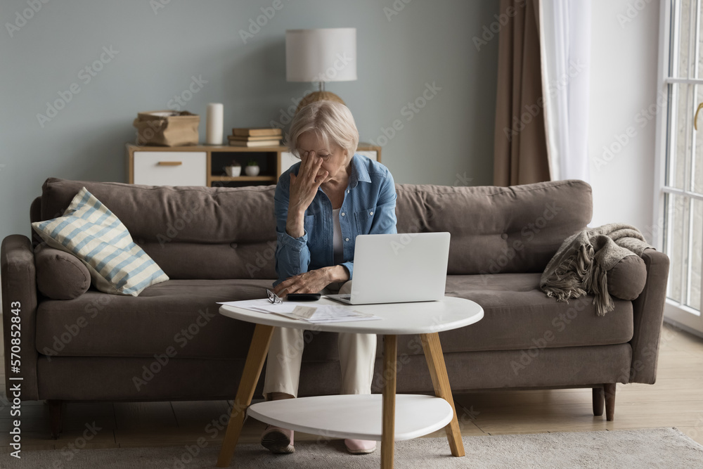 Frustrated desperate older lady getting bad news, financial problems, money loss, sitting on home couch at laptop, calculator, touching head, face with closed eyes, feeling headache Frustrated desperate older lady getting bad news, financial problems, money loss, sitting on home couch at laptop, calculator, touching head, face with closed eyes, feeling headache