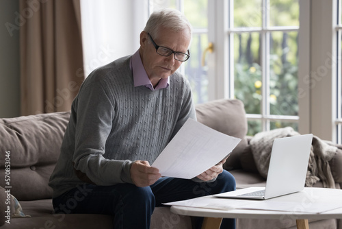 Focused elderly retired accountant man in glasses reviewing paper bill at laptop, paying loan, insurance, tax fees, reading document, letter, doing domestic household paperwork, sitting couch at home