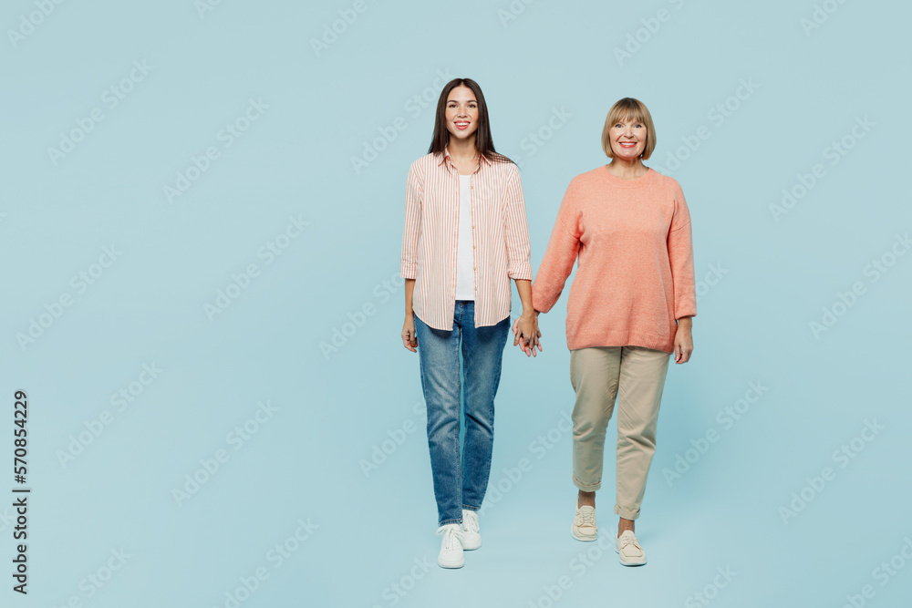 Full body smiling happy fun elder parent mom with young adult daughter two women together wearing casual clothes hold hands walk go look camera isolated on plain blue background. Family day concept. Full body smiling happy fun elder parent mom with young adult daughter two women together wearing casual clothes hold hands walk go look camera isolated on plain blue background. Family day concept.