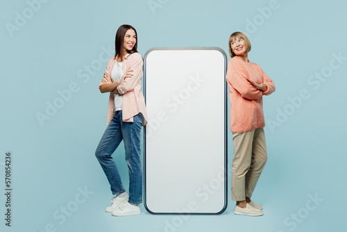 Full body side view elder parent mom with young adult daughter two women together in casual clothes big huge blank screen area mobile cell phone isolated on plain blue background Family day concept.