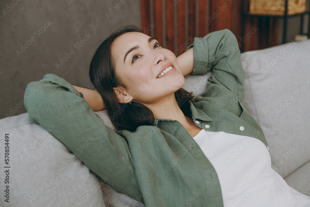 Side view close up young woman of Asian ethnicity wear casual clothes hold hands behind neck in reverie mood sits on grey sofa stay at home hotel flat rest relax spend free spare time in living room.