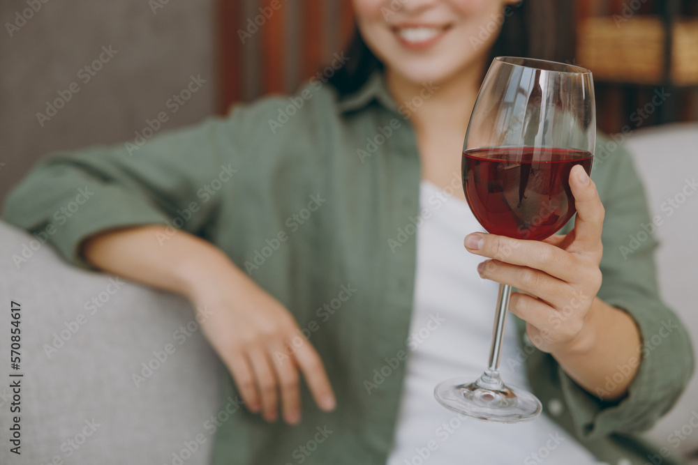 Cropped close up smiling cheerful fun oung woman wearing casual clothes drink red wine sits on grey sofa couch stay at home hotel flat rest relax spend free spare time in living room. Focus on glass.