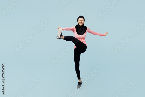 Full body fun young asian muslim fitness trainer sporty woman wear pink abaya hijab spend time in home gym raise up leg stretch isolated on plain blue background studio Workout sport fit abs concept