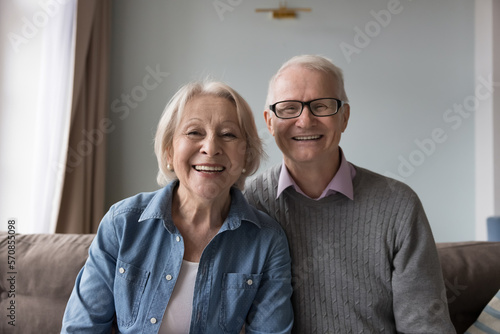 Happy older retired husband and wife sitting on home couch, looking at camera, talking on video call, smiling, laughing, enjoying family conversation. Cheerful elderly couple head shot portrait
