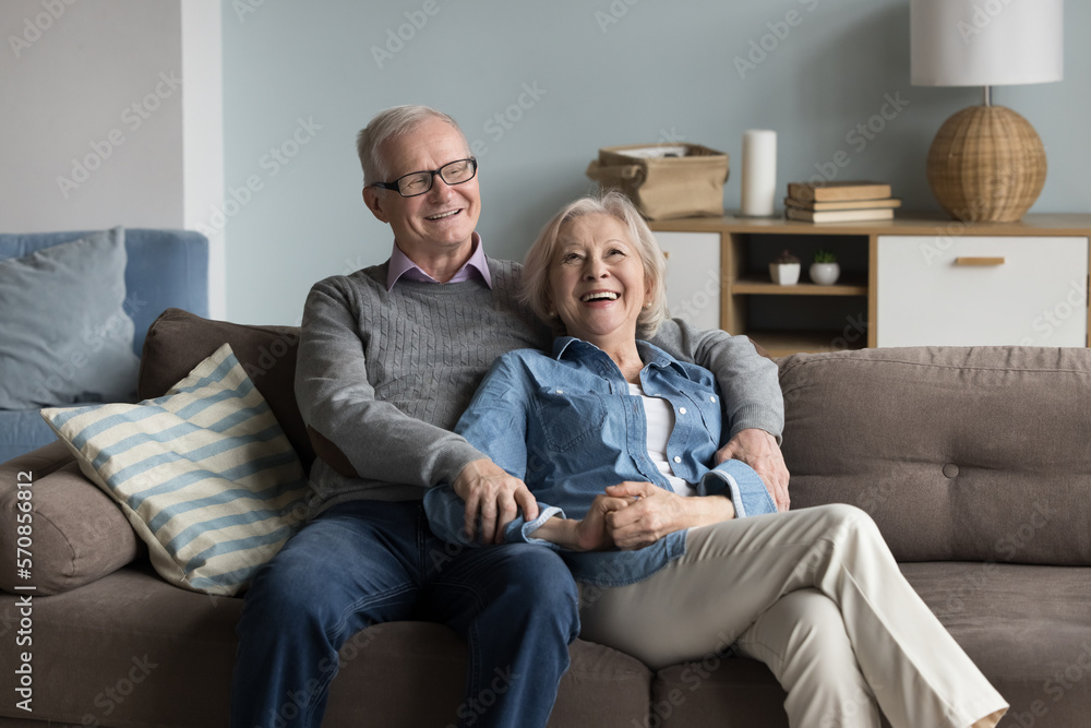 Cheerful relaxed older married couple resting on home couch, hugging with affectionate, talking, smiling, laughing, planning retirement, enjoying leisure together