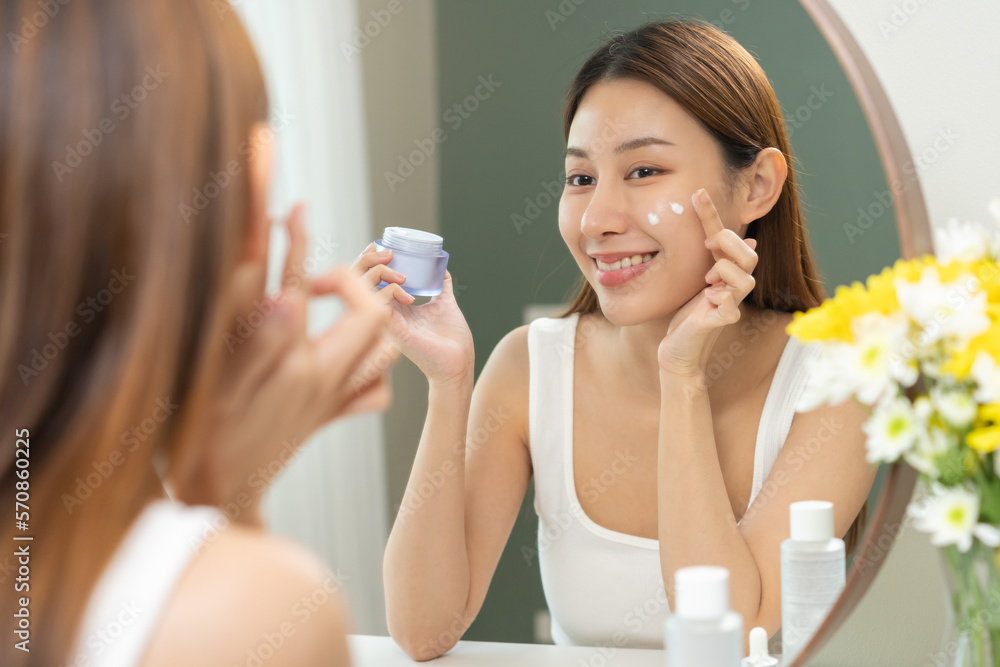 Fresh skin, beautiful smile of asian young woman, girl looking at mirror, hand applying moisturizer lotion on her face, putting cream treatment before makeup cosmetic routine at home. Facial Beauty. Fresh skin, beautiful smile of asian young woman, girl looking at mirror, hand applying moisturizer lotion on her face, putting cream treatment before makeup cosmetic routine at home. Facial Beauty.