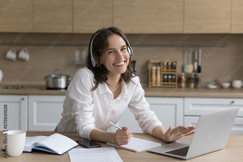 Portrait of happy beautiful young female student in wireless headphones sitting at home kitchen table, using laptop, notebook, writing notes, enjoying elearning process, looking at camera, smiling Portrait of happy beautiful young female student in wireless headphones sitting at home kitchen table, using laptop, notebook, writing notes, enjoying elearning process, looking at camera, smiling