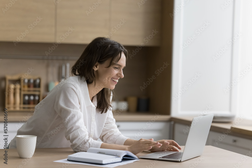 Happy young freelancer girl using laptop at home workplace table, typing, smiling, laughing at open notebook, talking on video call. Student enjoying remote learning, studying ea home Happy young freelancer girl using laptop at home workplace table, typing, smiling, laughing at open notebook, talking on video call. Student enjoying remote learning, studying ea home