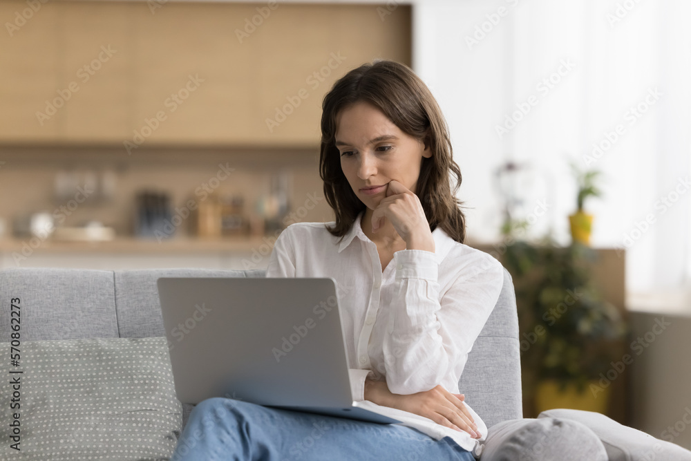 Serious thoughtful freelance employee girl using online app on laptop for job communication, working at computer, sitting on couch at home, reading email message, article, touching chin, thinking Serious thoughtful freelance employee girl using online app on laptop for job communication, working at computer, sitting on couch at home, reading email message, article, touching chin, thinking