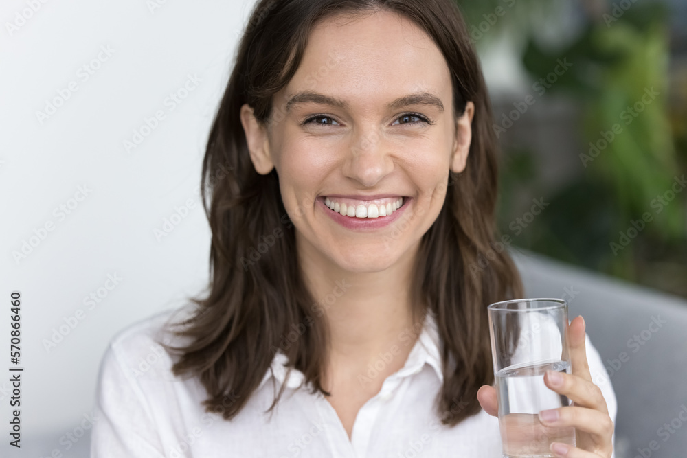 Happy pretty young adult girl drinking natural fresh water, holding glass, looking at camera with toothy smile, laughing, keeping healthy aqua balance, diet, lifestyle. Headshot home portrait Happy pretty young adult girl drinking natural fresh water, holding glass, looking at camera with toothy smile, laughing, keeping healthy aqua balance, diet, lifestyle. Headshot home portrait
