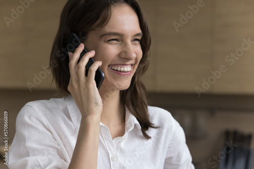 Happy young freelancer woman talking on mobile phone at home, enjoying telephone call, phone communication, speaking with toothy smile, laughing, looking away