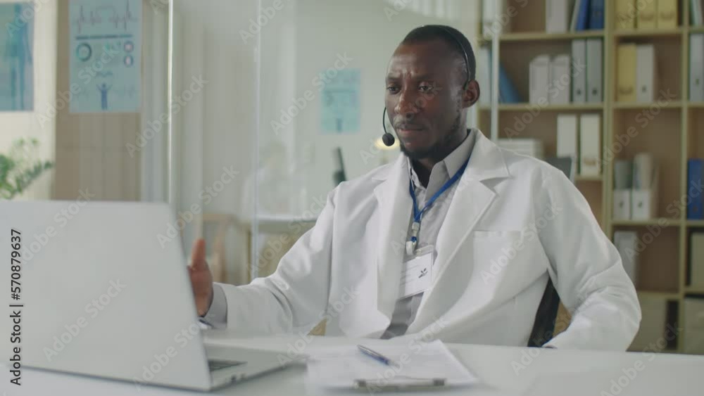 Medium shot of black male doctor in headset and white coat sitting at desk in clinic and speaking on video call on laptop while giving online consultation to patient Medium shot of black male doctor in headset and white coat sitting at desk in clinic and speaking on video call on laptop while giving online consultation to patient