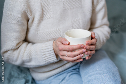 Young woman in white sweater and blue jeans sitting on the blue sofa and holding in her hands white cup of tee or coffee.