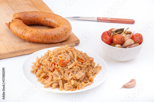 Stewed cabbage - bigos in a white porcelain plate against the background of a circle of smoked sausage and pickled vegetables on a light background photo