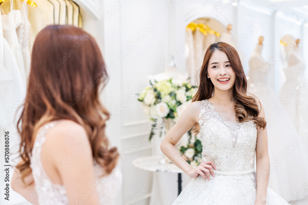 Young asian woman smiling and trying on wedding dress in a shop, asian bride look the mirror for beauty, Young female check beautiful reflection with happy smile for marriage, luxury or fashion Young asian woman smiling and trying on wedding dress in a shop, asian bride look the mirror for beauty, Young female check beautiful reflection with happy smile for marriage, luxury or fashion