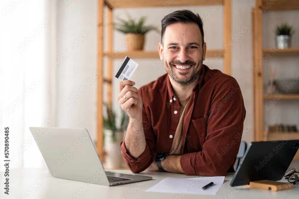 Confident businessman holding in hand showing credit card while sitting in home office. Caucasian businessman advertising banking services, loan or deposits, cashback for purchases from home concept. Confident businessman holding in hand showing credit card while sitting in home office. Caucasian businessman advertising banking services, loan or deposits, cashback for purchases from home concept.