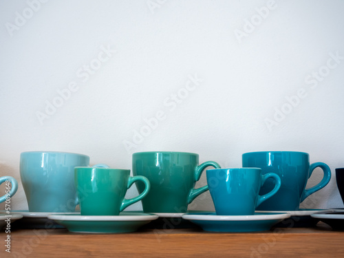 Set of big and small blue green ton of coffee mugs, ceramic cups with saucers stand in a row on wooden shelf isolated on white background with copy space.