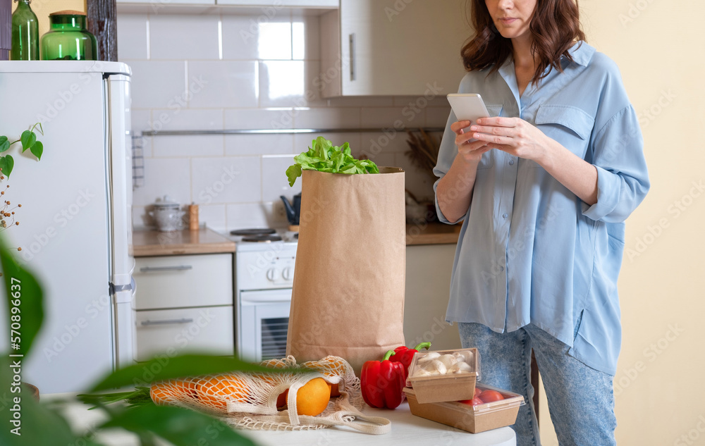 Young woman checking her fresh groceries delivery ordered from internet. Fresh organic vegetables, greens and fruits. Kitchen interior.  Food delivery concept