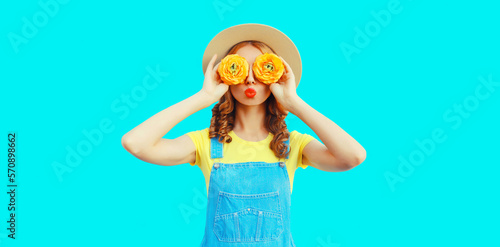 Summer portrait of happy young woman covering her eyes with flowers as binoculars looking for something wearing round straw hat, jumpsuit on blue background