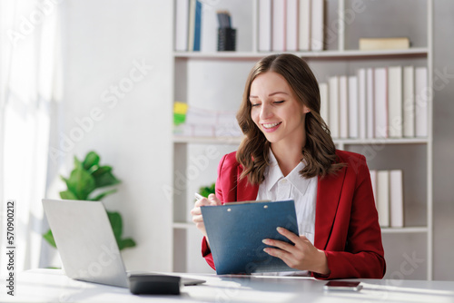 Concept of business working, Businesswoman wearing red suite sitting while writing and checking business analytic document  report  with computer laptop, smartphone on desk in her workstation.