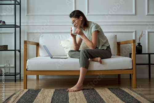 Young woman using laptop, sitting at home on sofa and drinking coffee, remote work, work at home